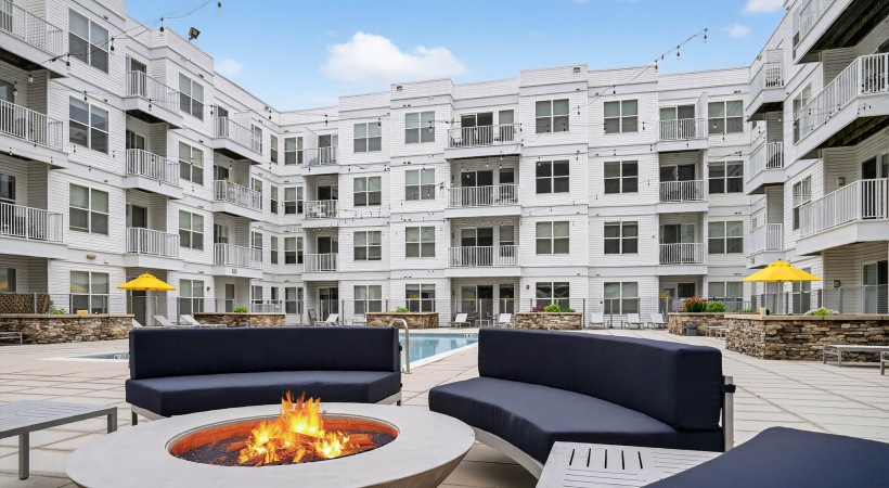 Fire pit with four cushioned couches surrounding, with view of swimming pool and buildings in background.